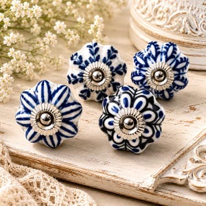 May include: Four ceramic drawer knobs with a scalloped, flower-like shape. Each knob features a unique blue and white hand-painted pattern and a central silver-toned metal hardware piece with a fluted, circular base and a smooth, rounded centre.