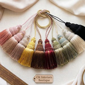 Puede incluir: Una colección de borlas decorativas en varios colores, incluyendo rosa, dorado, marrón y negro. Cada borla presenta un diseño detallado y está conectada a un cordón. La imagen incluye una regla de madera y una etiqueta que dice "OK Interiors Boutique".