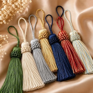 May include: A selection of decorative tassels in shades of green, cream, silver, gold, navy, and red. Each tassel has a looped top and a decorative knot, with long, flowing strands. The tassels are displayed on a satin-like fabric, creating a luxurious feel.