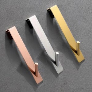 May include: Three modern metal wall hooks in copper, silver, and gold. Each hook has a rectangular backplate and a cylindrical hook extending from the bottom. The hooks are arranged diagonally against a gray background.