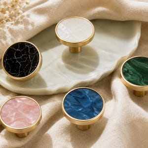 Könnte beinhalten: Eine Sammlung von fünf dekorativen Schrankknöpfen mit goldfarbenen Basen. Die Knöpfe haben runde Oberteile in verschiedenen Farben und Mustern, darunter schwarzer Marmor, Weiß, Rosa, Blau und Grün. Die Knöpfe sind auf einer hellen Oberfläche angeordnet.