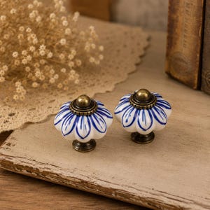 Puede incluir: Dos pomos de cerámica para armarios con base blanca y diseños de pétalos pintados a mano en azul. Cada pomo tiene una forma redondeada y estriada, rematada con un pequeño detalle metálico circular en tono latón antiguo.