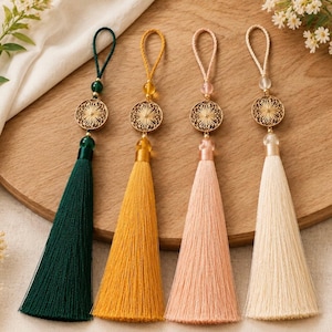 May include: Four decorative tassels in shades of green, yellow, pink, and cream. Each tassel has a gold-colored filigree disc and a loop for hanging. The tassels are displayed on a wooden surface, with floral accents.