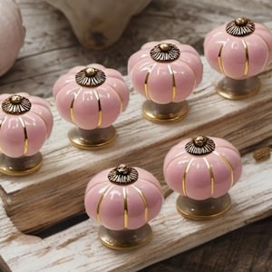 Puede incluir: Un conjunto de pomos de cerámica rosa con detalles dorados. Cada pomo tiene forma de calabaza, con rayas verticales doradas y una parte superior dorada decorativa. Se presentan sobre una superficie de madera, ideal para la decoración del hogar.