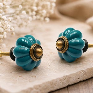 Puede incluir: Dos tiradores de cerámica turquesa con diseño floral. Cada tirador tiene un centro color latón y un tornillo dorado. Los tiradores se muestran sobre una superficie de piedra clara.