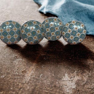 May include: Four decorative cabinet knobs with a patterned design in teal, gold, and white. The knobs are spherical and sit on a brown surface, with a blue cloth in the background.