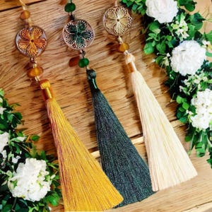 May include: Three decorative tassels with gold, green, and cream colored threads. Each tassel is attached to a round, beaded charm with a floral design.