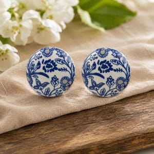 May include: Two round ceramic cabinet knobs with a blue and white floral design. The knobs are placed on a tan cloth, with white flowers and green leaves in the background. The knobs have a diameter of approximately 3.8 cm.