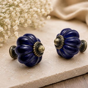 May include: Two navy blue ceramic cabinet knobs with a fluted design and antique brass hardware. The knobs are displayed on a light beige surface, with white flowers and a beige cloth in the background. Decorative hardware for furniture.