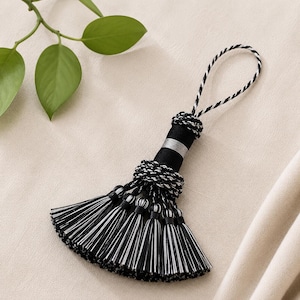 May include: A decorative tassel with a loop for hanging. The tassel features a black and white color scheme. The top section is black with a silver band, and the hanging loop is black and white. The bottom section has multiple black and white tassels.