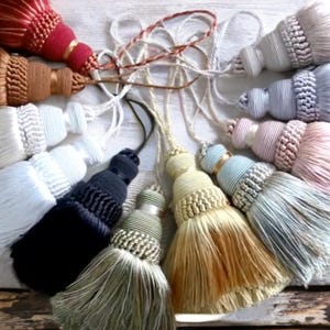 Könnte beinhalten: Eine Sammlung dekorativer Quasten in verschiedenen Farben, darunter Rot, Braun, Weiß, Schwarz und Pastelltöne. Jede Quaste hat ein detailliertes Design mit einer Schlaufenspitze und einem langen, gefransten Boden, ideal für Heimdekoration oder Bastelprojekte.