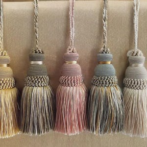 May include: Five decorative tassels in varying colors, including gold, pink, and gray. Each tassel features a detailed, layered design with a braided top and a flowing fringe. The tassels are suspended against a neutral background.