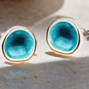 May include: A pair of cufflinks with a white base, a gold rim, and a turquoise center. The cufflinks have a unique, organic shape and a shiny, reflective surface. The background is a neutral, light color.