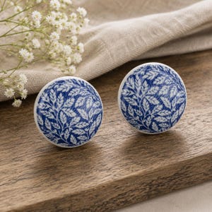Puede incluir: Dos pomos redondos de cerámica con un estampado de hojas azules y blancas. Los pomos están sobre una superficie de madera, con una ramita de flores blancas y un paño beige en el fondo. Estos pomos decorativos son perfectos para cualquier mueble.