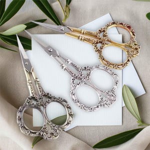 May include: Three pairs of decorative scissors with ornate floral designs. The scissors are silver, gold, and a combination of silver and gold.