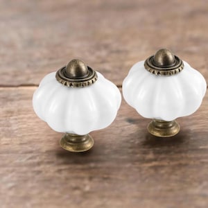May include: Two white ceramic cabinet knobs with a pumpkin-like shape. Each knob has a bronze-colored base and a bronze-colored cap. The knobs are set against a wooden background.
