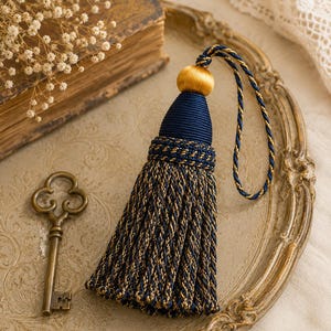 May include: A decorative tassel with a navy blue and gold design. The tassel features a gold ball at the top, navy blue fabric, and a braided gold and navy blue cord. The tassel is on a decorative tray with a vintage key.