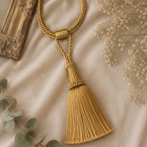 May include: A gold-coloured curtain tieback featuring a twisted rope cord and a decorative tassel. The tassel consists of a woven, structured top section that transitions into a dense, straight fringe. The cord forms a loop with a central knot.