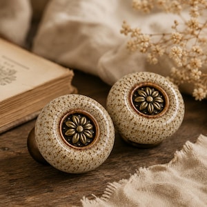 May include: Two round ceramic cabinet knobs featuring a speckled beige and brown finish. Each knob has a central circular inlay made of antique brass-toned metal, embossed with a detailed flower design with multiple petals and a small centre dot.