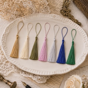 May include: A set of seven decorative tassels in gold, cream, olive green, light pink, silver, royal blue, and emerald green. Each tassel features a matching braided cord loop at the top and a straight, silky fringe extending from a central knot.