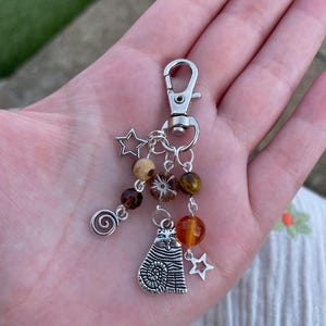 May include: A silver keychain with a lobster clasp, featuring a cat charm, star charms, and brown and orange beads. The keychain includes a spiral charm and a variety of bead shapes. The beads are in shades of brown and orange.