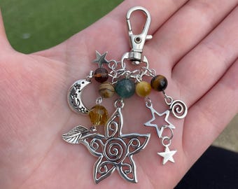 May include: Silver-toned keychain with a lobster clasp, adorned with a star-shaped charm, a crescent moon, and various star charms. The keychain also features a collection of colorful beads, including brown, green, and yellow.