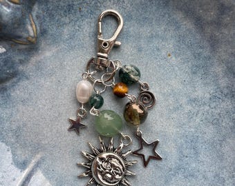 May include: A silver keychain with a collection of beads and charms. The keychain features a sun and moon charm, star charms, and various green, brown, and white beads. The keychain is attached to a silver clasp.