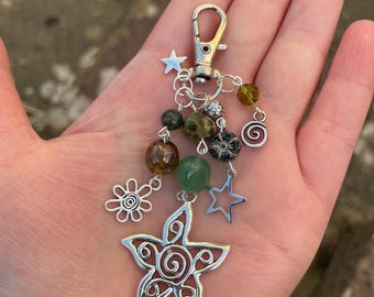 May include: A silver-toned keychain with a lobster clasp, adorned with a mix of beads in green, brown, and yellow hues. Decorative charms include a star, a flower, and a large openwork star, creating a whimsical accessory.
