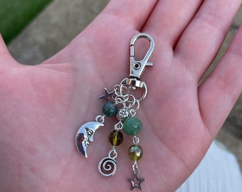May include: A silver-toned keychain with celestial charms. The keychain features a crescent moon, stars, and a spiral charm, along with green and brown beads. The clasp is a lobster claw style.