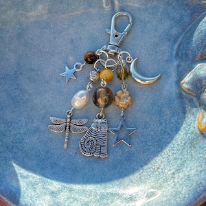 May include: A silver keychain with a selection of charms, such as a dragonfly, cat, star, and crescent moon. The keychain also incorporates beads in brown, yellow, and green, plus a pearl. The clasp is silver, and the design has a whimsical aesthetic.