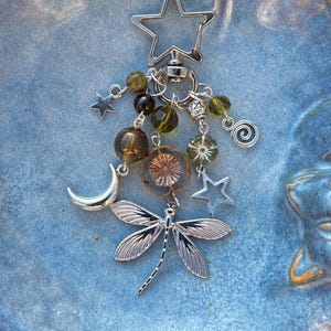 May include: A silver keychain with celestial charms. The keychain features a star-shaped clasp, a dragonfly pendant, and dangling beads in shades of green and brown. Additional charms include a crescent moon, stars, and a spiral.