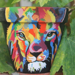 May include: A colorful painted terracotta flower pot with a lion's face design. The lion's face is painted in a variety of bright colors, including red, yellow, blue, green, and purple.