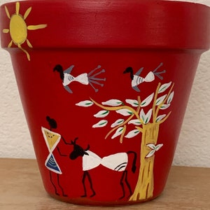 May include: A red painted terracotta flower pot with a yellow sun, birds, and figures. The pot features a tree and animal design in white, black, and yellow. The pot is sitting on a wooden surface.