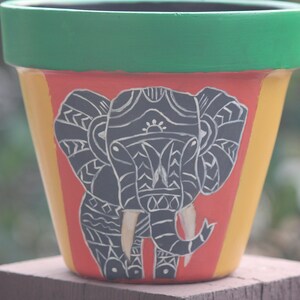 May include: A terracotta flower pot painted with a black and white elephant design. The pot is decorated with green, orange, and yellow stripes.