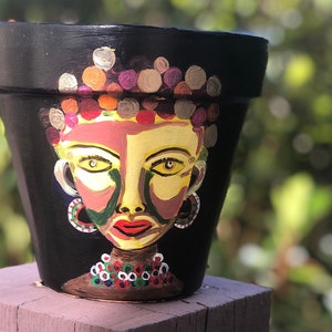 May include: A black terracotta flower pot painted with a colorful portrait of a person. The person has yellow eyes, red lips, and a brown necklace with white and green accents.