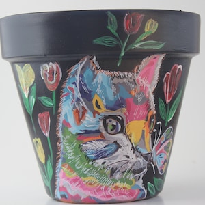 May include: A black terracotta planter with a hand-painted design of a colorful cat with a butterfly. The cat's fur is painted in shades of blue, green, pink, and yellow. The butterfly is painted in shades of pink and purple.