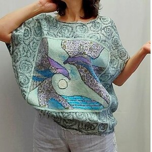 May include: A light blue, short-sleeved top with a unique design. The top features a central panel with a depiction of two stylized figures in shades of purple, blue, and green. The fabric has a textured appearance, and the sleeves are loose-fitting.