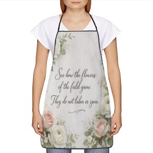 May include: White apron with a floral design featuring pink and white roses, green leaves, and the text "See how the flowers of the field grow. They do not labor or spin." The apron has a black trim and neck strap.