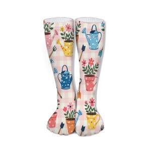 Könnte beinhalten: Kniestrümpfe mit einem rosa-weiß karierten Muster. Die Socken sind mit farbenfrohen Illustrationen von Gießkannen, Blumentöpfen und Gartengeräten verziert. Das Design umfasst rosa, gelbe, blaue und orangefarbene Elemente.
