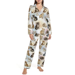 May include: A light-coloured pajama set with a repeating pattern of cat illustrations in brown, grey, and cream. The set includes a button-down top with a V-neck and long sleeves, and matching straight-leg trousers.