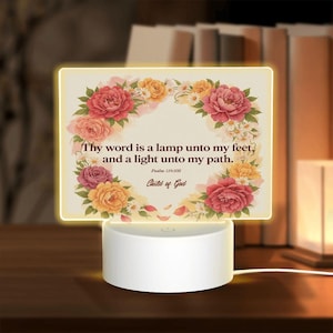 May include: A rectangular acrylic night light with a white circular base. The acrylic panel features a floral wreath of pink, yellow, and white flowers surrounding the text: Thy word is a lamp unto my feet, and a light unto my path. Psalm 119:105 Child of God.