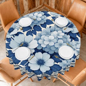 May include: A round tablecloth featuring a floral design in shades of blue and white. The pattern includes large flowers and leaves, with a cream-colored background. White plates and gold utensils are set on the table, ready for a meal.