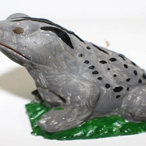 Puede incluir: Una figurita de rana de cerámica gris con manchas y rayas negras. La rana está sentada sobre una base verde.