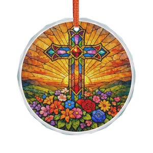 May include: Round ornament with a stained-glass design featuring a cross with a red gem at the center. The cross is set against a sunset backdrop with colorful flowers at the base. An orange ribbon is attached.
