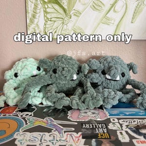 May include: Three crocheted plush toys in shades of green. The toys are shaped like monsters with large eyes and toothy grins. The toys are sitting on a laptop with stickers. The text "digital pattern only @jfs.art" is visible in the image.