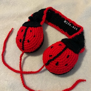 May include: Crocheted ladybug earmuffs in red and black. The earmuffs have a black band and red ear covers with black spots. The text "@jfs.art" is on the band. The ear covers are connected by a red cord.