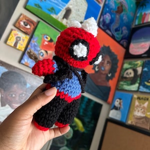 May include: A crocheted red and black character with white eyes and horns. The character is wearing a blue and black outfit.