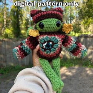 May include: A crocheted green frog wearing a brown, red, and green sweater with yellow flower accents. The frog is holding a sign that says "digital pattern only" and the text "@jfs.art" is visible on the frog's sweater.