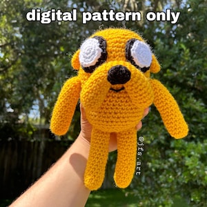 May include: A crocheted yellow dog with white eyes and a black nose. The dog is being held by a hand. The text "digital pattern only" is written above the dog. The text "@jfs.art" is written below the dog.