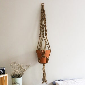 May include: A hanging macrame plant holder with a terracotta pot. The macrame is made of brown rope and is suspended from a metal ring. The pot is empty. The holder is hanging on a white wall.